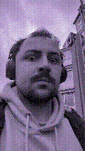 a selfie of a white person, wearing either headphones or a helmet, making an odd expression. the photo is edited to be colorized into a monochrome purple-ish color palette.